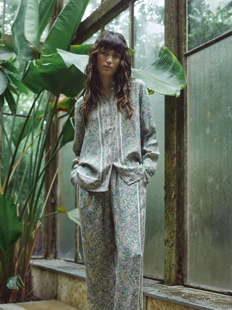 Person standing in a greenhouse with large plants and glass walls.