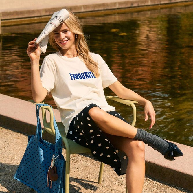 Woman sitting by a pond holding a blue bag, wearing a white t-shirt with text and dark polka dot skirt.